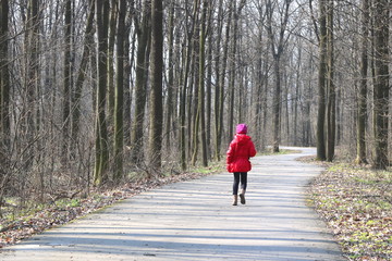 Little girl in red jacket and red hat walks in spring forest among trees on asphalt road