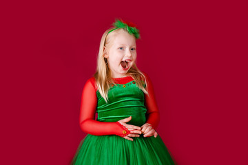 Little blonde girl in a christmas and new year green costume having fun and sending kisses on red background. Space for text. Dreams come true Merry Christmas