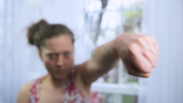 Slow motion of aggressive brunette woman in shallow depth of field blurred background punching boxing at camera with angry furious expression, defending herself from danger, fighting at home 