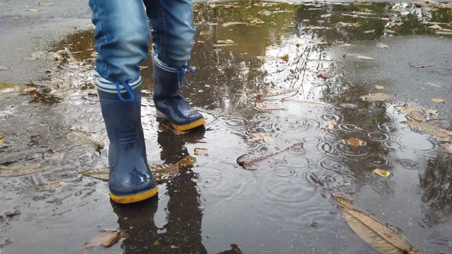 Boy with blue rain boots playing in a puddle with autumn trees reflection.