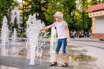 Obraz premium Little caucasian boy in hat playing and having fun with water in fountain in the sunny summer park