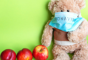 A soft toy bear in a protective medical mask with the inscription "coronavirus", sitting on a green background, next to apples. The concept of quarantine of schools and kindergartens, home education.