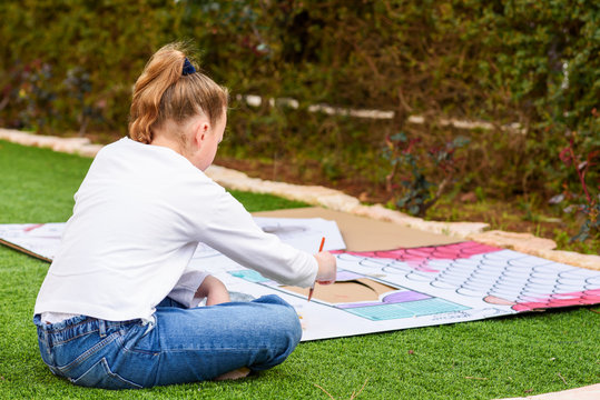 Child play outdoors. Back view teenager girl paints a paper house template with colorful watercolors on the grass in the garden.