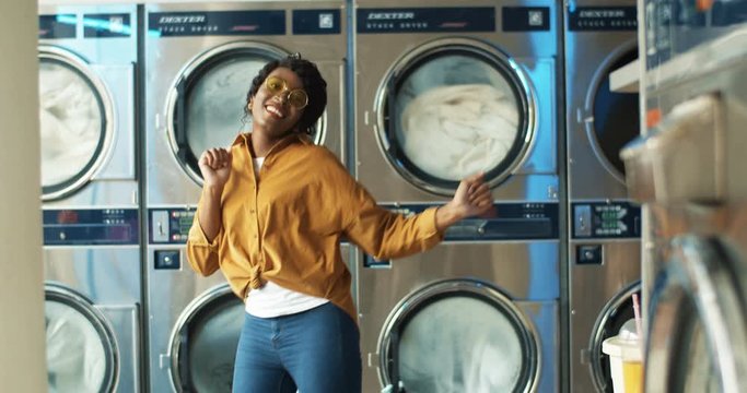 Beautiful African American cheerful woman in stylish outfit having fun and dancing in laundry service room while machines washing on background. Girl resting while machines working in washhouse.