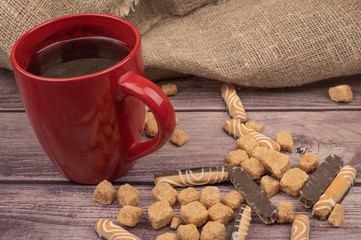 A red ceramic tea mug, cookie sticks with white icing and chocolate , and pieces of brown cane sugar on a wooden background. Close up.