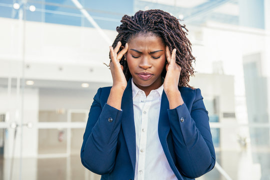 Stressed tired female office employee suffering from headache. Young black business woman with closed eyes and pain face standing outside, touching head and temples. Fatigue or sickness concept