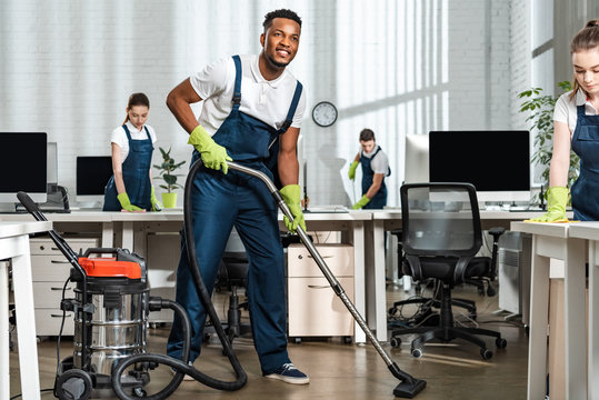 smiling african american cleaner vacuuming floor near team of multicultural colleagues