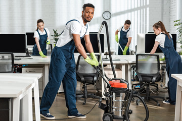 smiling african american cleaner moving vacuum cleaner near team of multicultural colleagues