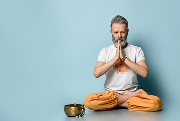 Mature gray-haired man in dhoti clothes. Praying, sitting on floor in lotus pose on blue...