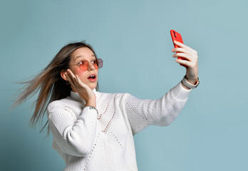 Teenager in orange sunglasses, watch, bracelet and sweater. She taking selfie, surprised, posing on blue background. Close up