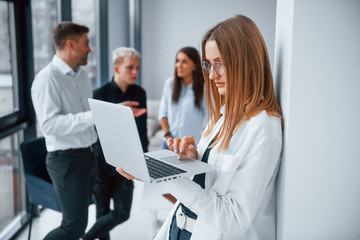 Blonde with laptop stands in front of young successful team that working and communicating together indoors in office