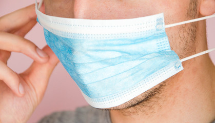 A man in a gray T-shirt on a pink background puts on a medical blue mask on his face. Virus protection. COVID-19