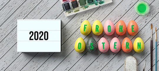 easter eggs with message HAPPY EASTER in German and lightbox with number 2020 surrounded by water color boxes on wooden background