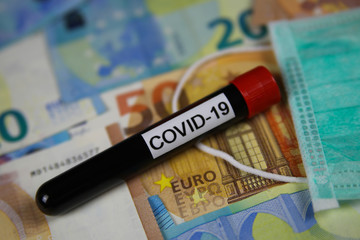 Corona virus uncertain consequential costs concept symbol: closeup of isolated blood sample vial on euro paper money bank notes und surgical mask