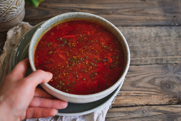 Red borsch in a bowl of gray clay. Tomato soup, borsch. Wooden background Healthy vegetarian and vegan food. Woman holds in hand a bowl and spoon. Food and hands in the frame.