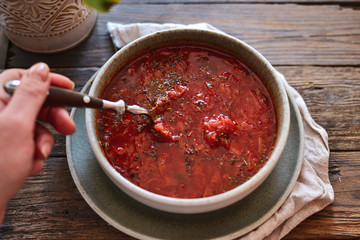 Red borsch in a bowl of gray clay. Tomato soup, borsch. Wooden background Healthy vegetarian and vegan food. Woman holds in hand a bowl and spoon. Food and hands in the frame.