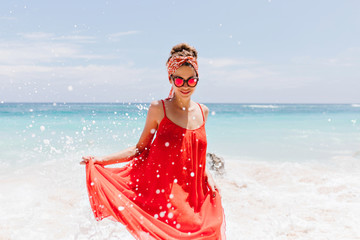 Attractive girl in trendy red dress posing on nature background. Outdoor shot of winsome female model in romantic attire standing near ocean.