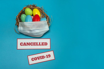 The concept of canceling events due to the Covid-19 coronavirus pandemic. A medical mask, a nest with colorful Easter eggs and warning signs on a light blue background. Free space.