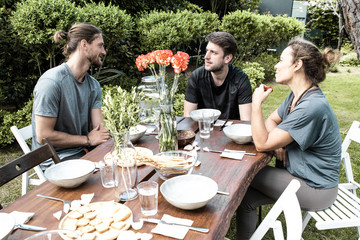 Smiling people talking in backyard. Cheerful young male and female friends sitting at table with organic meal and talking outdoor. Gathering together concept