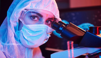 Nurse in mask and white uniform sitting in neon lighted laboratory with computer, microscope and medical equipment searching for Coronavirus vaccine
