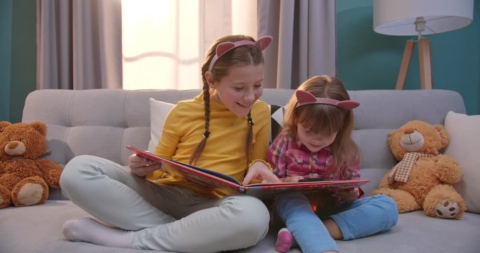 Caucasian cute small kids in funny ears sitting on couch and reading book in cozy living room. Pretty little sisters read fairytale. Girls exploring textbook with magnifying glass at home.