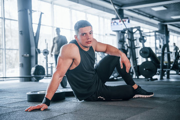Fototapeta na wymiar Strong young man in sportive clothes have fitness day in the gym, sitting and taking a break