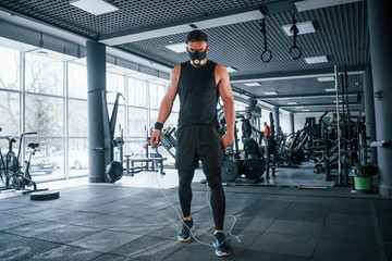 Strong young man in sportive clothes and scary mask doing exercise by using jump rope in gym