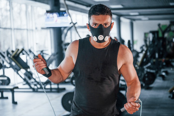Fototapeta na wymiar Strong young man in sportive clothes and scary mask standing with jump rope in gym