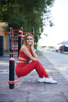 Young blond woman, wearing red fitness outfit sitting on horizontal bar on sports playground. Healthy active life concept. Close-up picture of girl in process of doing exercises outside in the city.