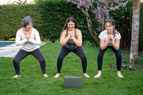 Mother, Grandmother And Teen Daughter Practicing Fitness Lesson Online Outdoors In Garden At Quarantine Isolation Period During Coronavirus Pandemic. Family Doing Sport Together At Home Via Skype