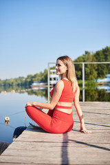 Fototapeta na wymiar Young blond woman, wearing red fitness outfit sitting on wooden pier dock in summer morning. Healthy active life concept. Portrait of girl resting after training exercise outside in the city.