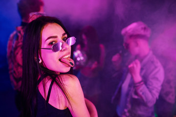 Young girl holding drug pills on togue in front of young people that having fun in night club with...