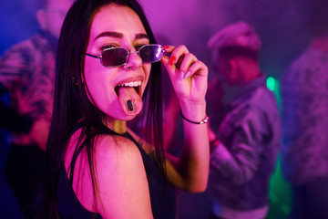 Young girl holding drug pills on togue in front of young people that having fun in night club with...