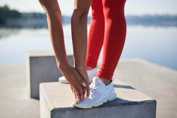 Young woman, wearing red fitness outfit and white sneakers stretching legs on concrete platform by city lake. Healthy active life concept. Close-up picture of legs and hands in process of training.