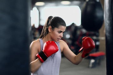 Athletic young brunette woman in sportswear and red boxing gloves trains bumps on a punching bag in...