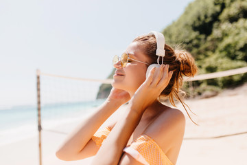 Close-up portrait of joyful tanned woman relaxing with favorite music at beach. Outdoor shot of smiling female model in headphones spending time at resort.