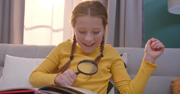Caucasian cute teen child reading book and sitting on sofa in cozy living room. Pretty little kid read fairytale. Girl exploring textbook with magnifying glass at home. Small curious reader concept.