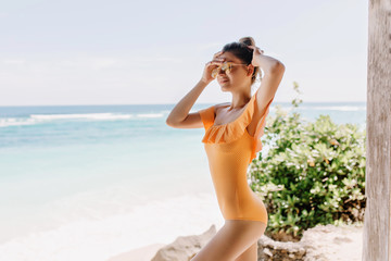 Fototapeta na wymiar Graceful white female model in yellow clothes posing on ocean background. Slim caucasian girl in orange swimsuit chilling near sea with eyes closed.