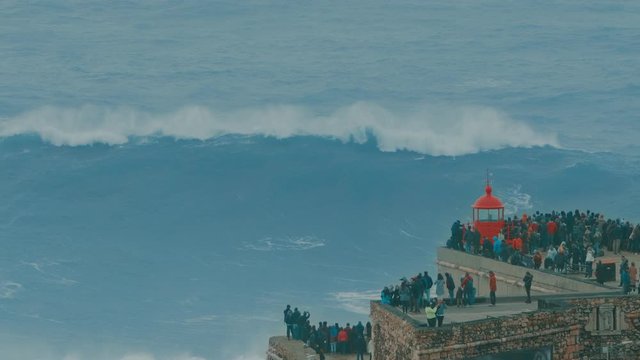 NAZARE, PORTUGAL - DECEMBER 22, 2019: Amazing shot of a big wave coming towards the red lighthouse on a cliff in Nazare, Portugal. Popular tourist spot and travel destination for people who love extre