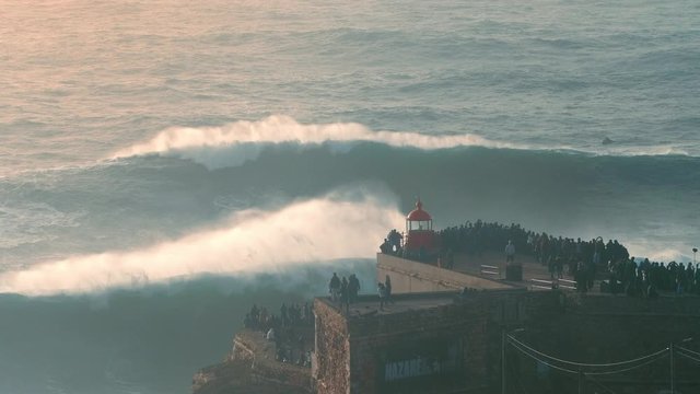 NAZARE, PORTUGAL - DECEMBER 22, 2019:  Amazing shot of a big wave coming towards the red lighthouse on a cliff in Nazare, Portugal.  Popular tourist spot and travel destination 