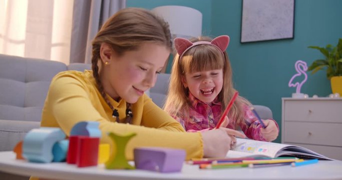 Cheerful small Caucasian sisters sitting at home alone and playing nice together. Cute kids coloring picture with colorful pencils in cozy room. Happy children play and talking indoor.