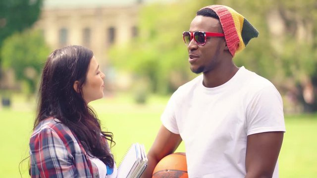 A young African American student playing street basketball with friends met his classmate who he likes, decided to chat with her while standing on a path in a campus campus holding a basketball.