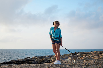 Young blond woman, wearing jeans shorts and turquoise blouse, standing at the beach by the sea, holding light grey white husky puppy with purple leash. Woman, walking with dog at seaside.