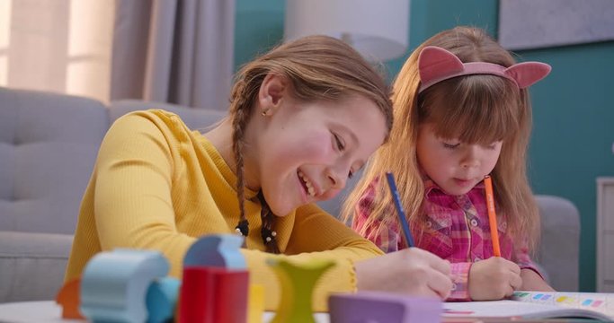 Close up of funny little Caucasian sisters staying at home alone and playing joyfully together. Nice kids coloring picture with colorful pencils in cozy room. Children having fun, play and chatting.