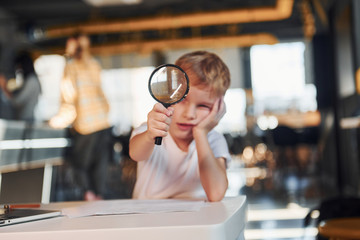 Smart child in casual clothes with laptop on table have fun with magnifying glass