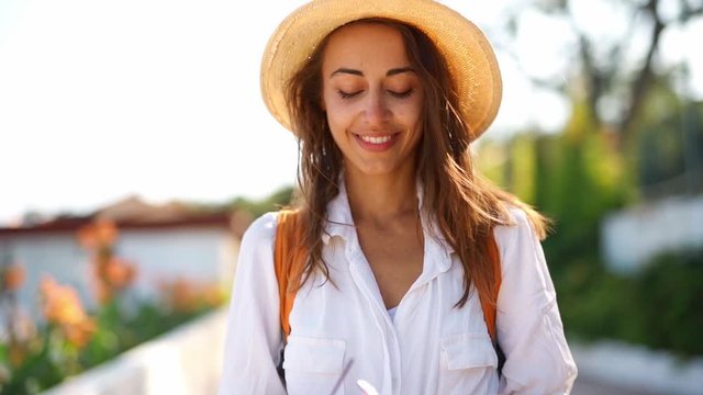 Young charming woman tourist in white shirt and straw sunhat pretty smiles, looking straight in camera and puts on sunglasses. 120 fps slow motion footage
