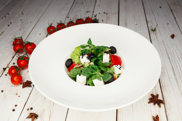 Greek salad of vegetables, cream cheese, olives and herbs served on a large white plate. Salad on a white wooden table.