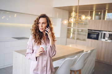 Beautiful young woman with curly hair drinking coffee at home in lazy weekend morning in cozy pink...