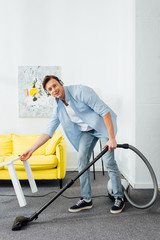 Smiling man in headphones looking at camera while cleaning carpet with vacuum cleaner in living room