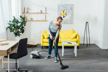 Fototapeta na wymiar Young woman in headphones cleaning carpet with vacuum cleaner in living room